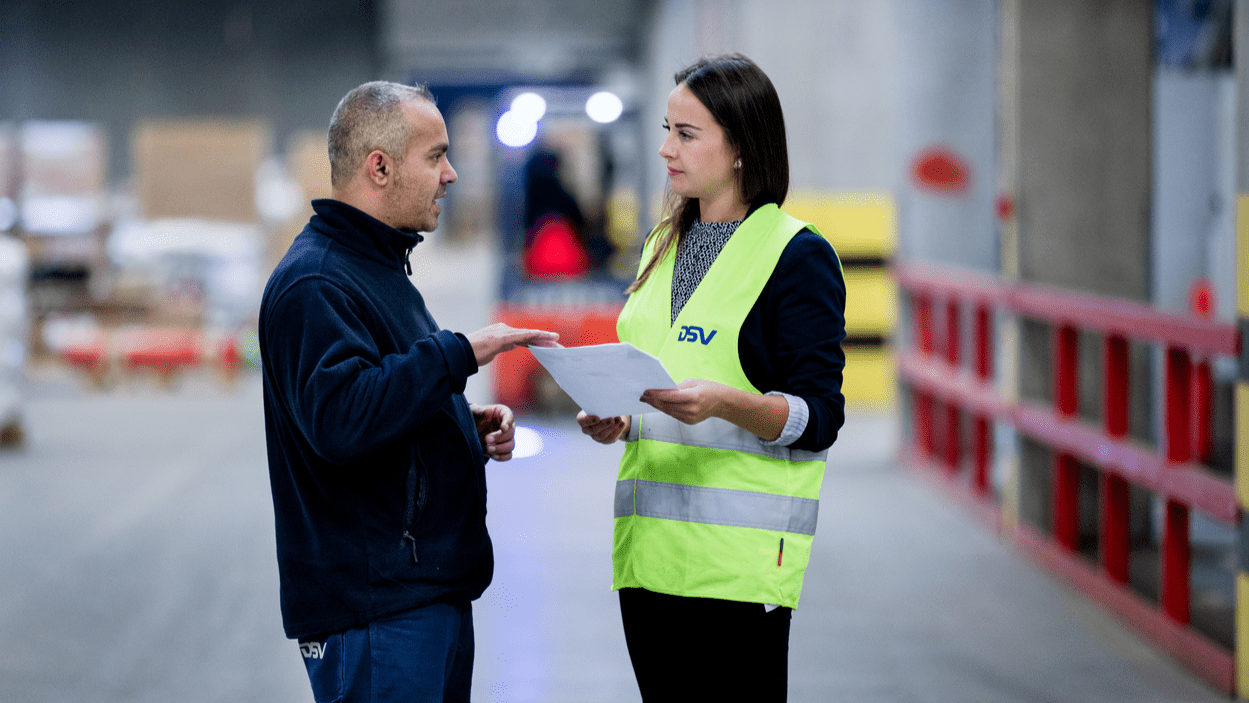 Mann i fleece og kvinne i refleksvest diskuterer mens truck kjører i bakgrunnen
